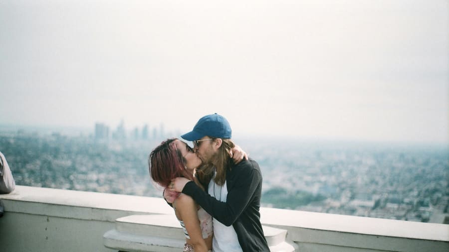 A woman and her companion sharing a kiss at a railing high above a hazy city skyline.