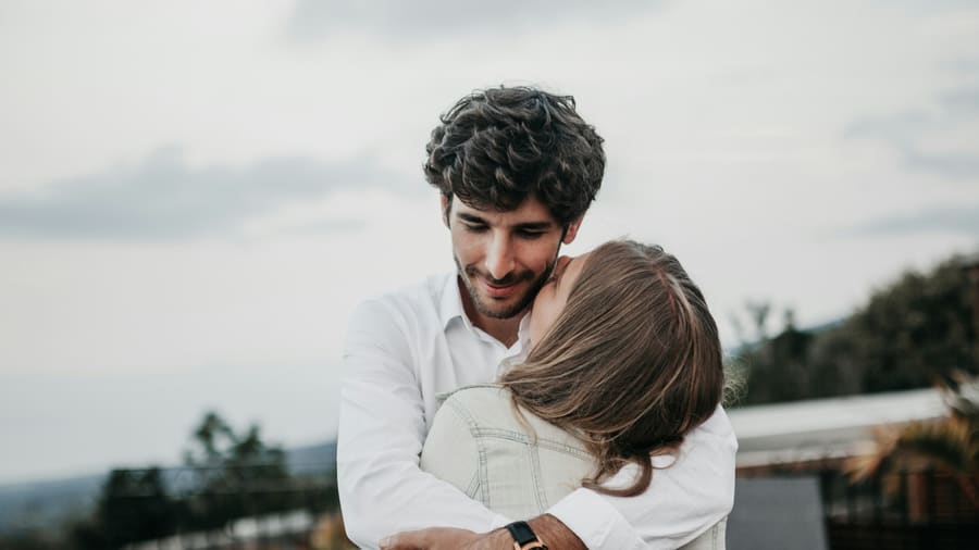 A woman resting her face against her companion's shoulder as they hug on a hillside overlook under an overcast sky.