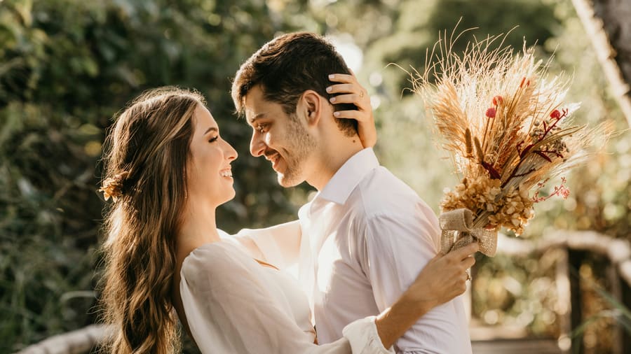 A woman and her companion smiling with their foreheads close together, a bouquet of dried flowers held beside them in soft daylight.