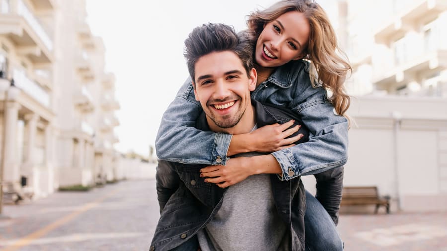 A woman laughing on her companion's back in a piggyback along a sunny street of pale buildings.