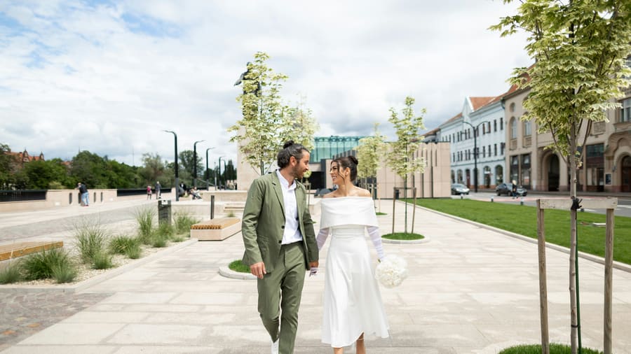 A woman in a white wedding dress walking hand in hand with her companion across a bright town square.