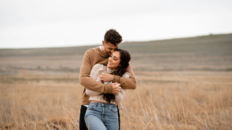 A woman smiling as her companion embraces her from behind in a wide field of tall dry grass.
