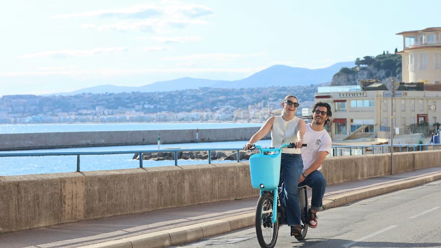 A woman and her companion riding a shared bicycle along a seafront promenade beside a blue bay.
