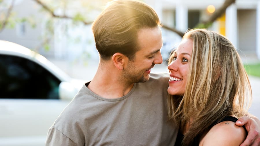 A woman and her companion laughing face to face on a sunlit street, arms around each other.