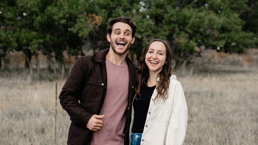 A woman and her companion standing side by side, both laughing, in a dry meadow in front of trees.